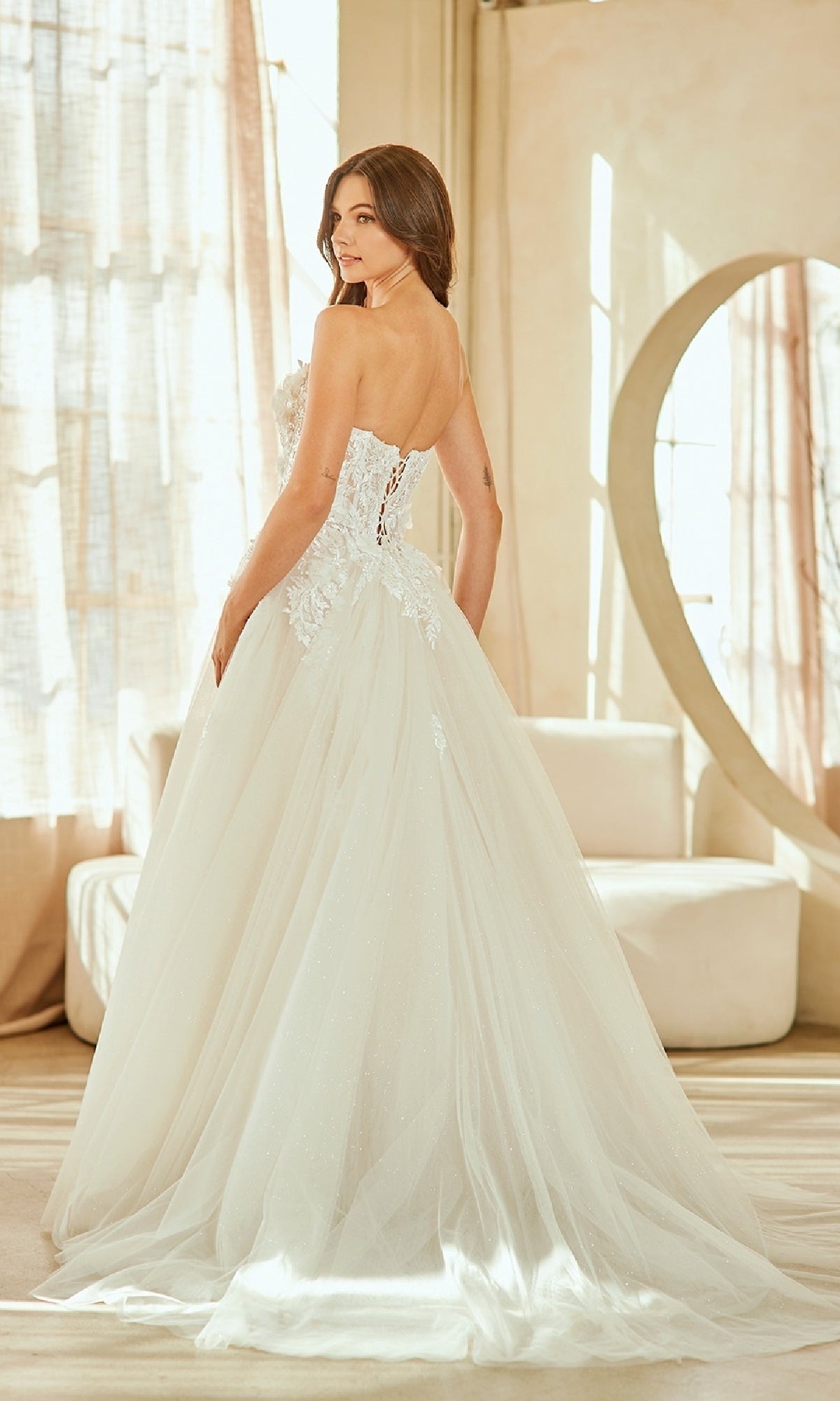 A woman stands indoors, looking over her shoulder in the Amelia Couture 2141 long wedding dress featuring a strapless ball gown silhouette and lace-up corset bodice.